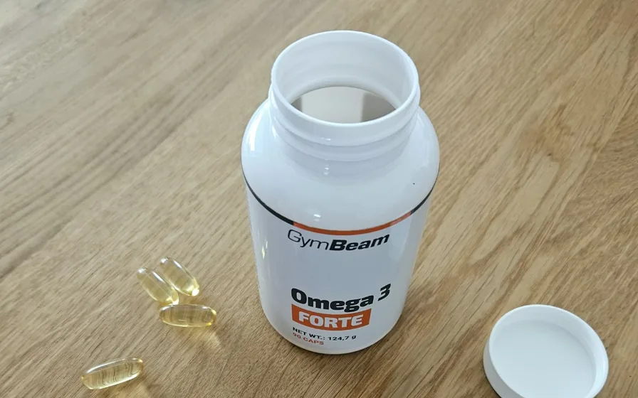 Geöffnete Dose GymBeam Omega 3 Forte mit Deckel und vier Weichkapseln auf einem Holztisch