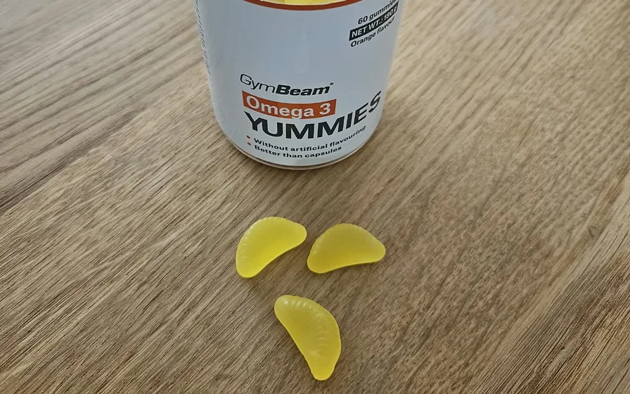Dose GymBeam Omega 3 Yummies mit drei gelben Fruchtgummis in Form von Orangenspalten auf einem Holztisch