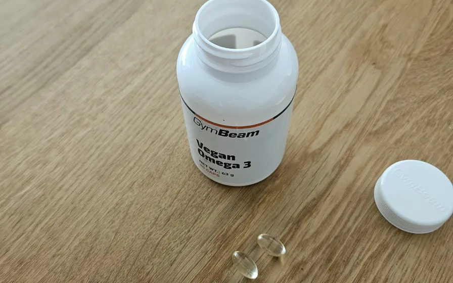 Geöffnete Dose GymBeam Vegan Omega 3 mit Deckel und zwei Weichkapseln auf einem Holztisch