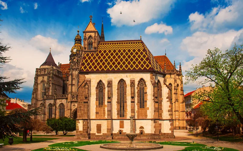 Gotische Kirchenbauten mit bunt gemusterten Ziegeldächern in Košice, davor ein Brunnen